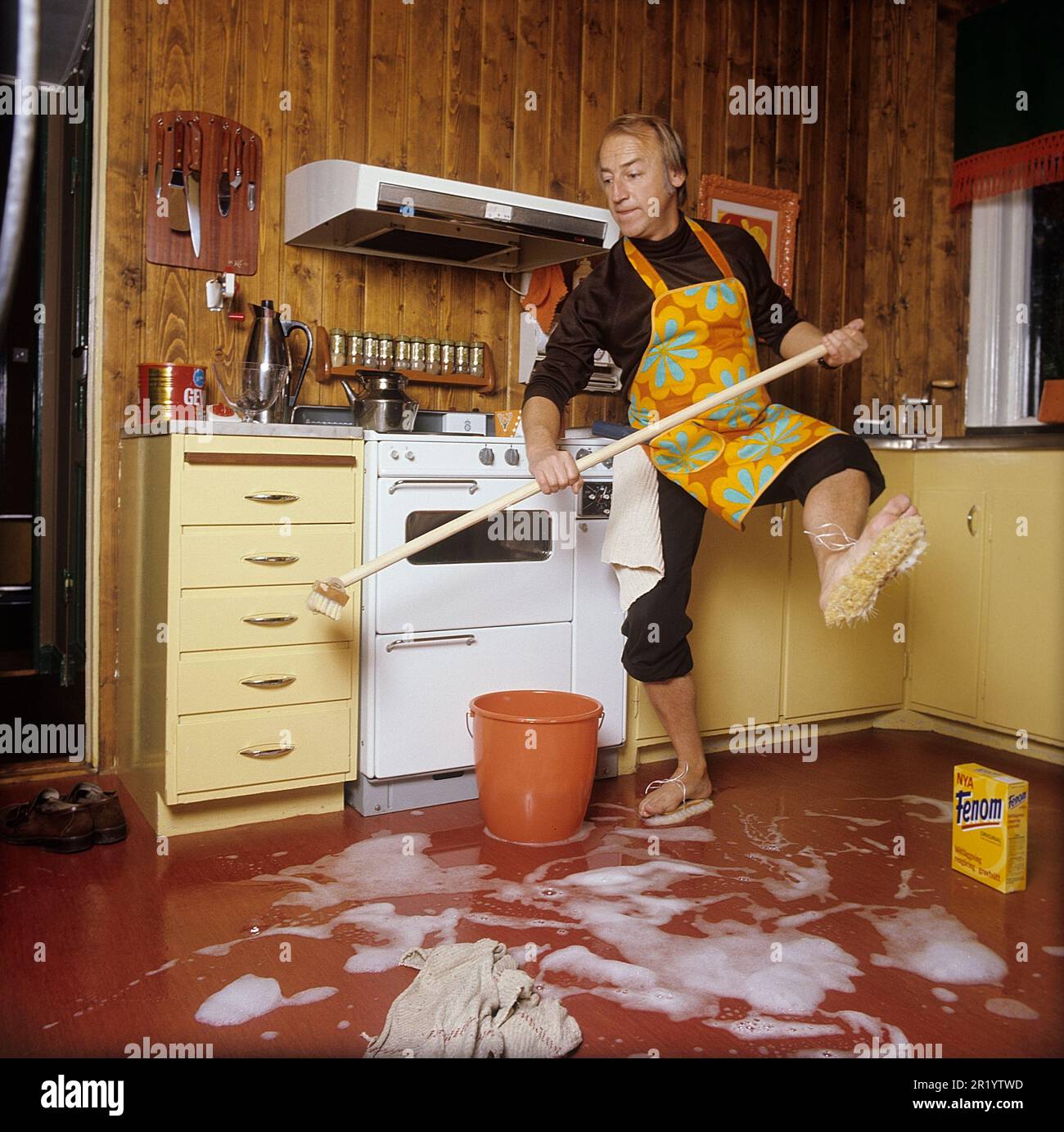 Jour de nettoyage en 1970s. Intérieur d'une cuisine où l'on voit un homme nettoyer le sol. Pour accélérer le travail, il a monté des brosses sur ses pieds. Il est acteur Stig Grybe 1928-2017. 4 octobre 1972. Kristoffersson Banque D'Images