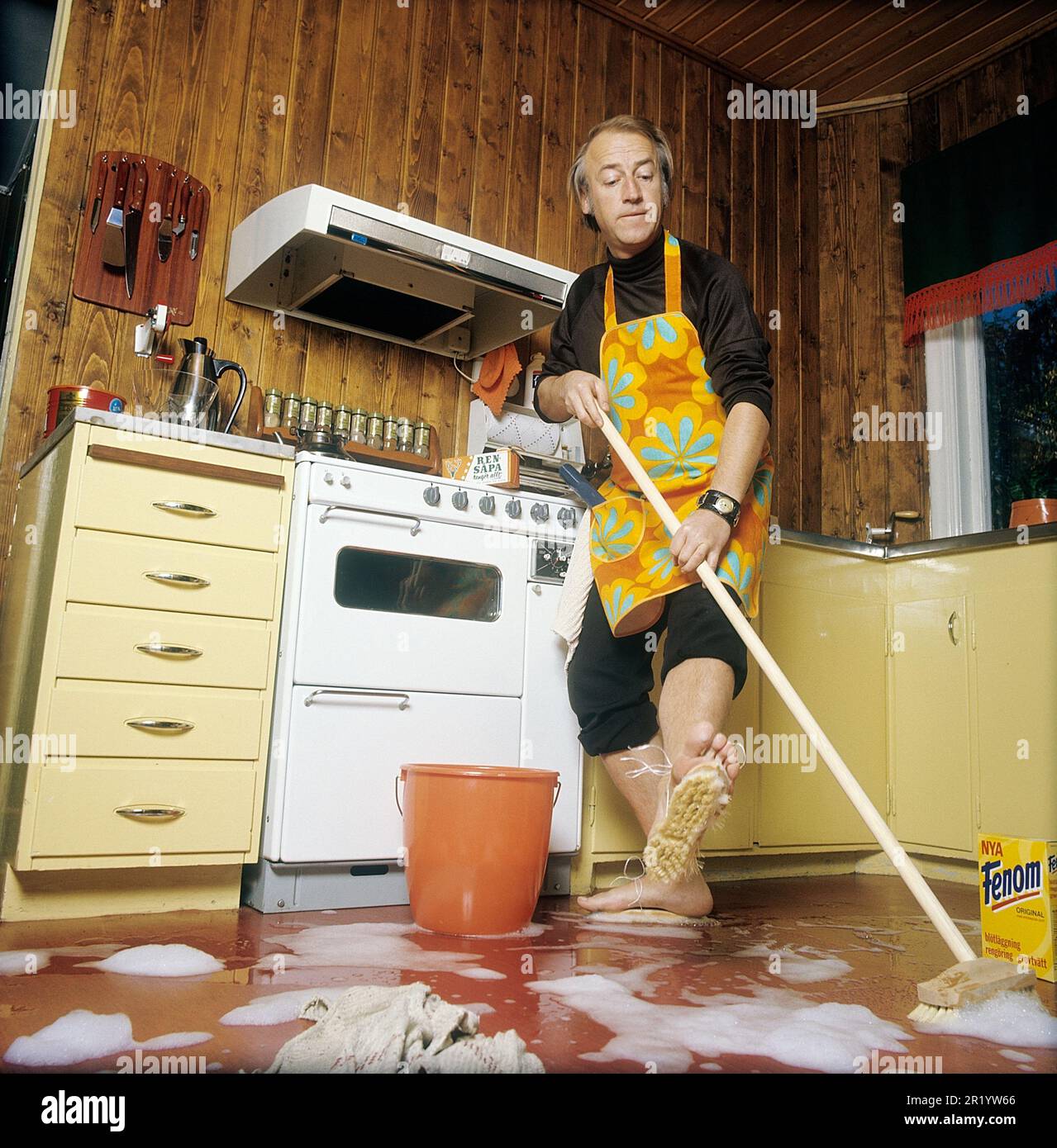 Jour de nettoyage en 1970s. Intérieur d'une cuisine où l'on voit un homme nettoyer le sol. Pour accélérer le travail, il a monté des brosses sur ses pieds. Il est acteur Stig Grybe 1928-2017. 4 octobre 1972. Kristoffersson Banque D'Images