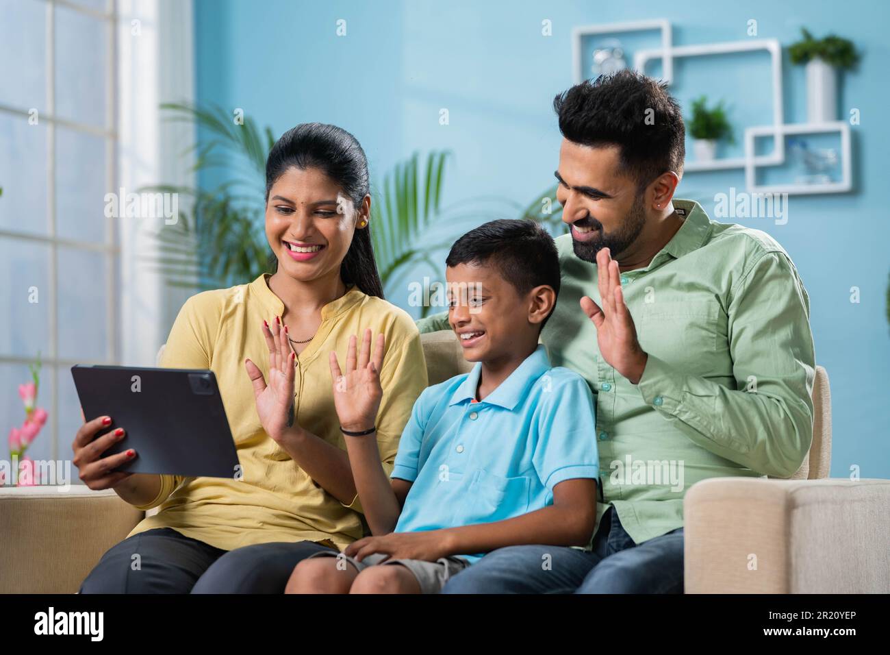 Couple indien souriant avec un enfant qui fait des appels vidéo sur une tablette numérique tout en étant assis sur un canapé à la maison - concept de technologie, de relation et de conne Banque D'Images