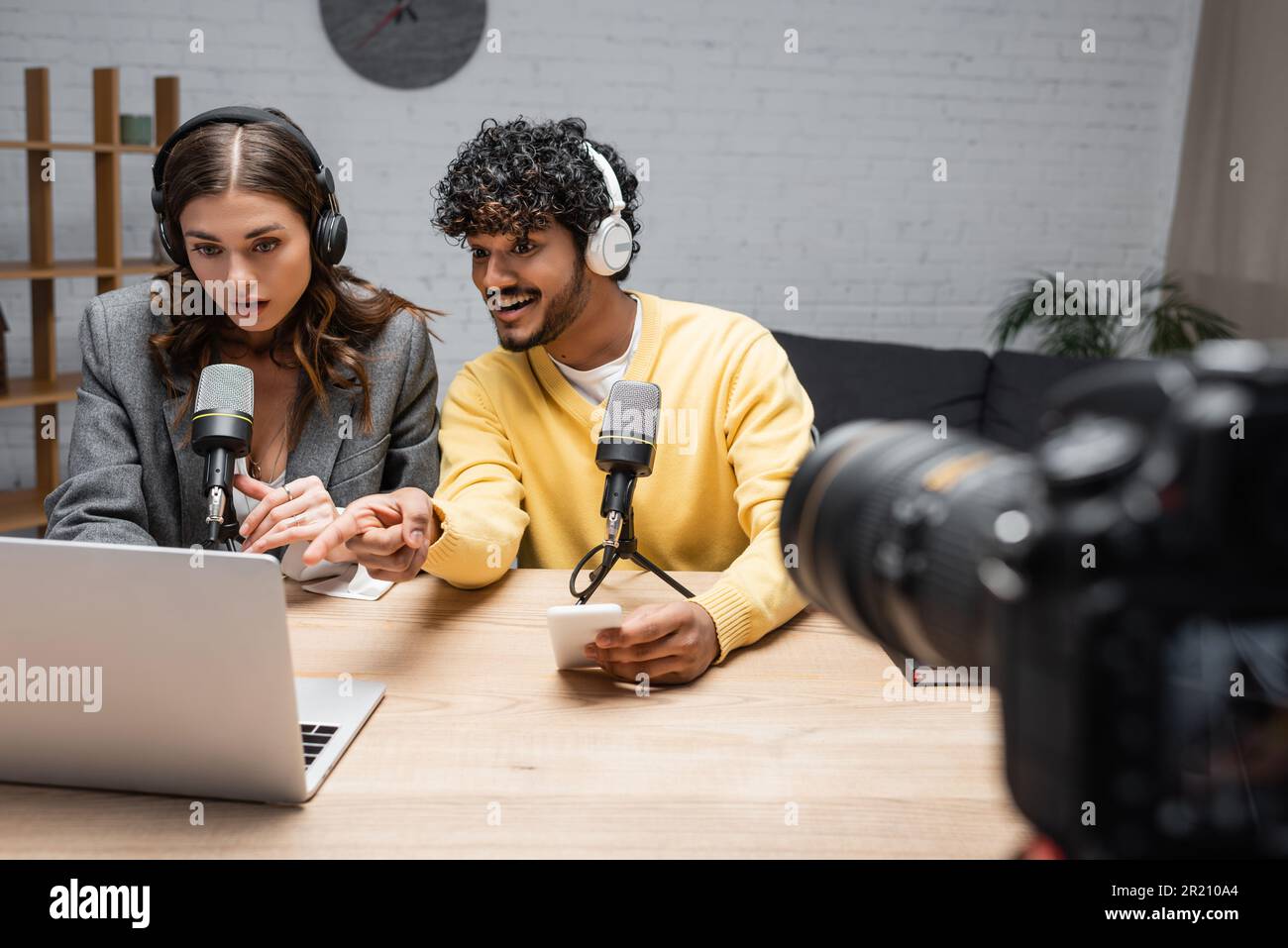 homme indien joyeux dans un casque et un cavalier jaune tenant un smartphone et pointant vers un ordinateur portable près d'un collègue brunette stupéfait et d'un appareil photo numérique sur blu Banque D'Images