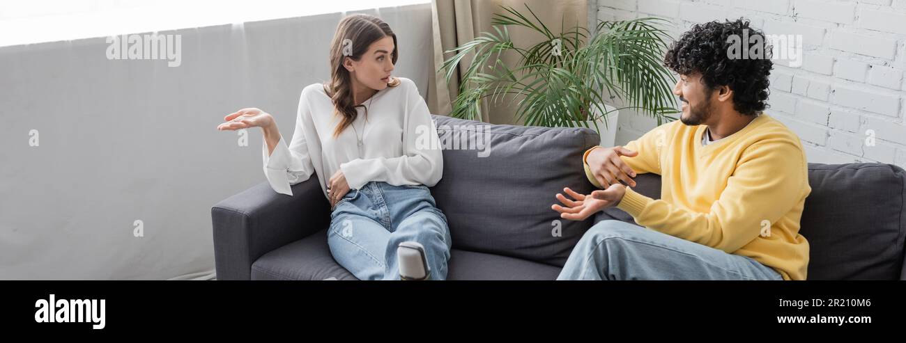 femme émotive brune en blouse blanche gestante en parlant à un jeune et mauriquement indien en cavalier jaune assis sur le canapé près de la plante verte à nouveau Banque D'Images
