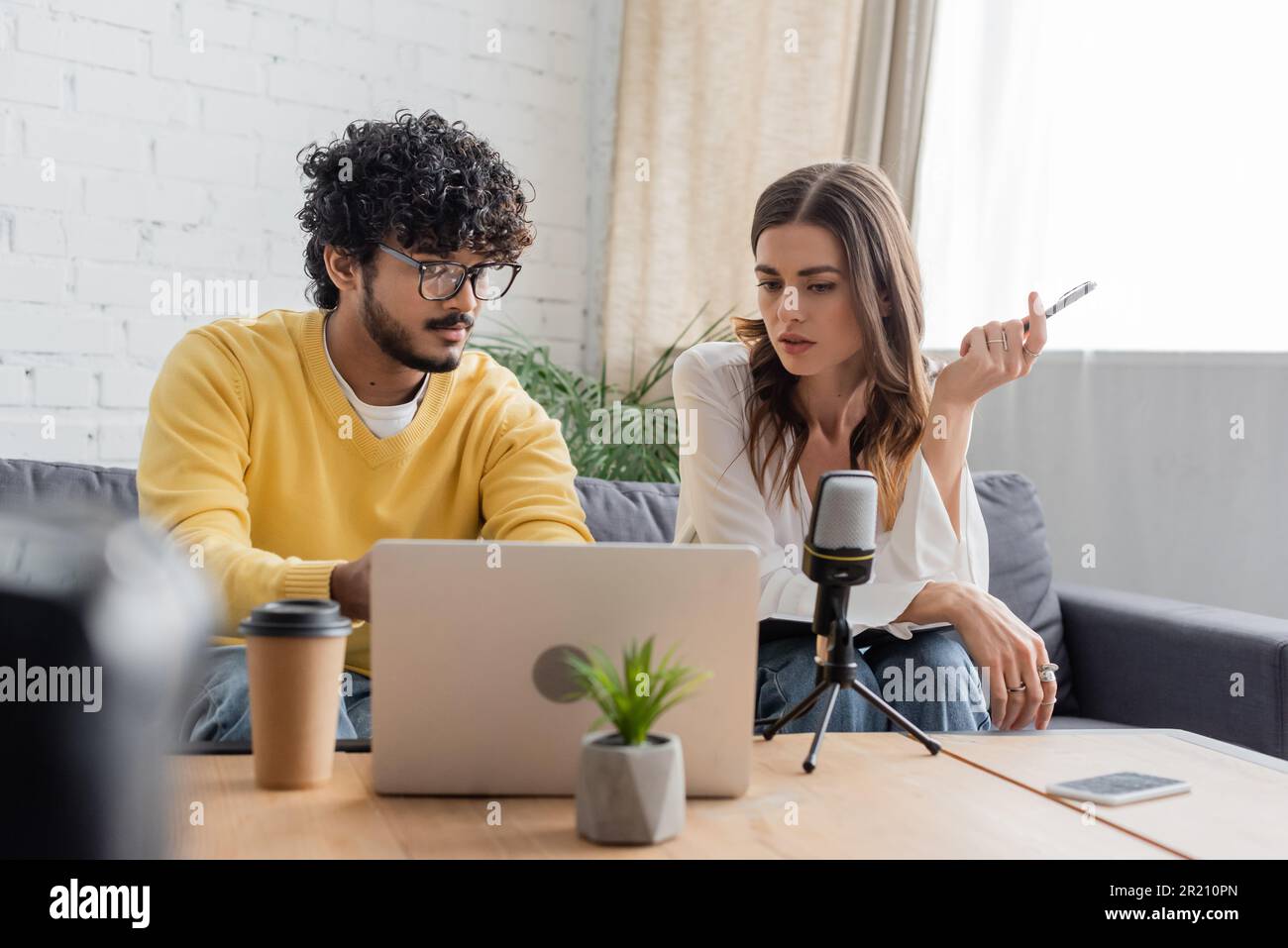 les diffuseurs interraciaux réfléchis qui regardent un ordinateur portable lors d'un appel vidéo près d'un microphone, d'un smartphone avec écran vierge, d'une boisson à emporter et d'un pot de fleurs Banque D'Images