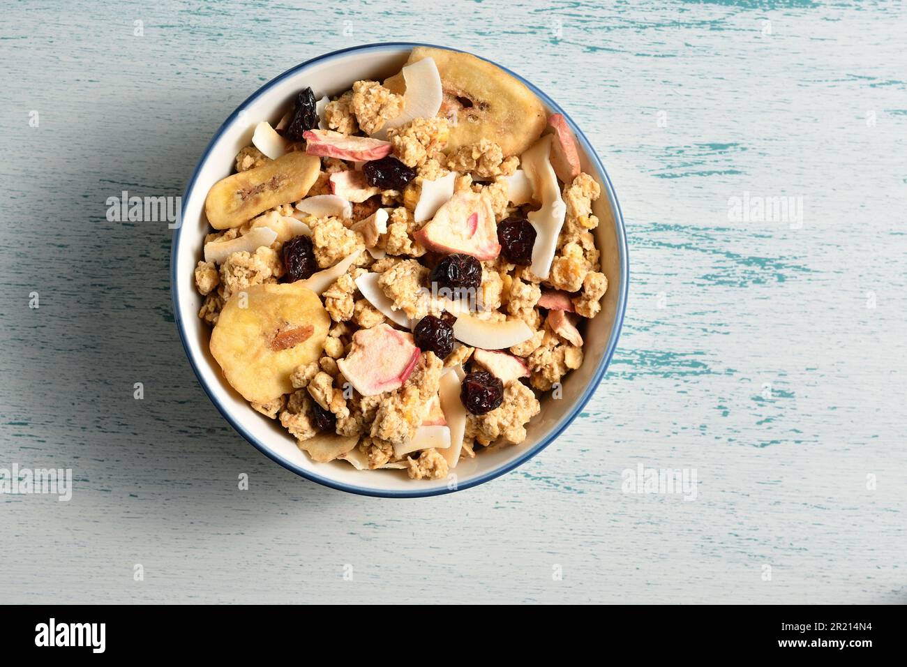 Bol de délicieux muesli de petit déjeuner avec noix de coco séchée, banane, pomme, canneberges, raisins secs sur un arrière-plan chic en bois. Banque D'Images