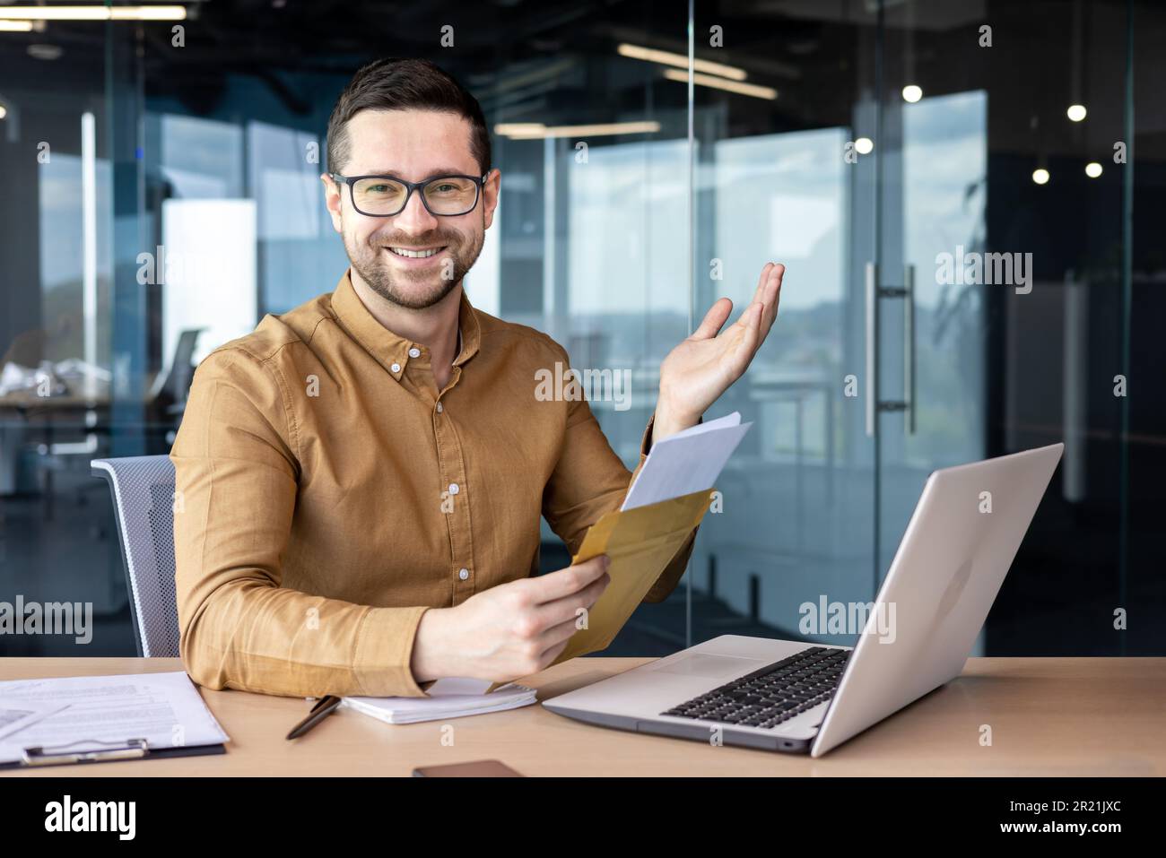 Portrait d'un homme assis au bureau et travaillant sur un ordinateur portable. A reçu et tient dans ses mains une enveloppe avec une lettre et des documents. Un look souriant Banque D'Images