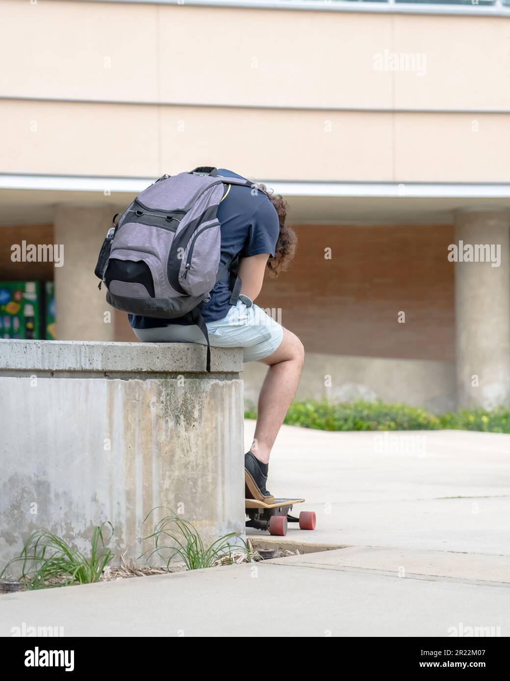 Un étudiant de l'université assis a chassés avec son skateboard électrique et son sac à dos, campus, et regardant son téléphone entre les classes. Banque D'Images