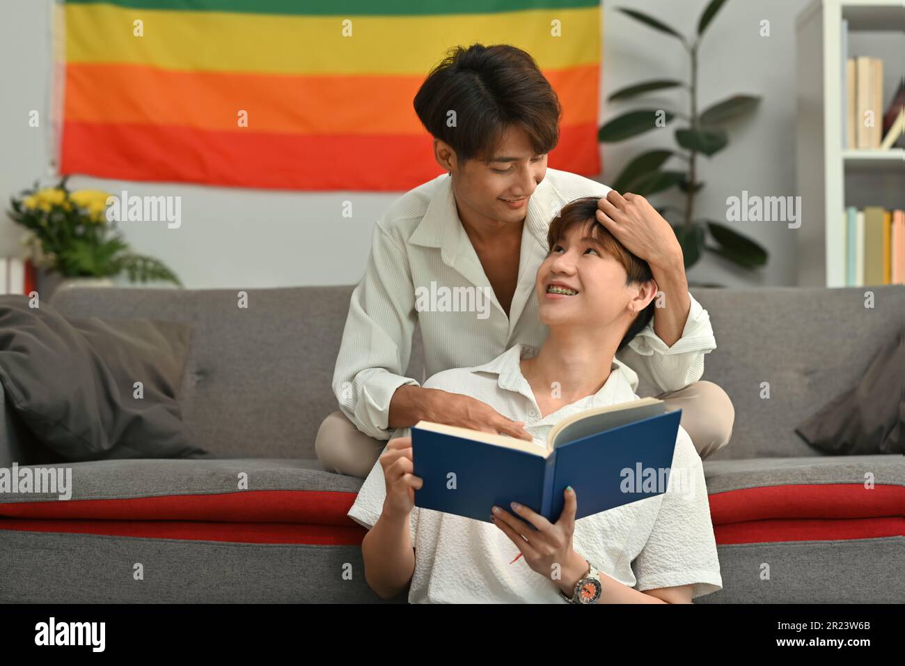 Un couple gay heureux lisant le livre sur le canapé à la maison, passant du temps ensemble le week-end. Les personnes LGBTQ vivent et aiment l'émotion Banque D'Images