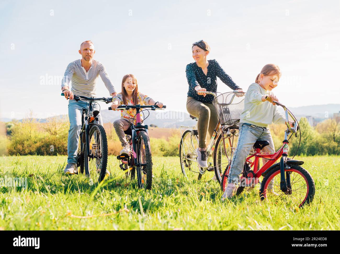 Père souriant et mère avec deux filles pendant l'été, vélo en plein air. Ils aiment la convivialité sur la prairie verte haute herbe. Bonne parenthoo Banque D'Images