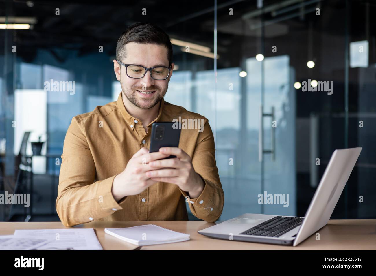 Un jeune travailleur de bureau souriant s'assoit à un bureau avec un ordinateur portable et écrit un message au téléphone, discute, vérifie le courrier, lit les nouvelles, fait une pause au travail Banque D'Images