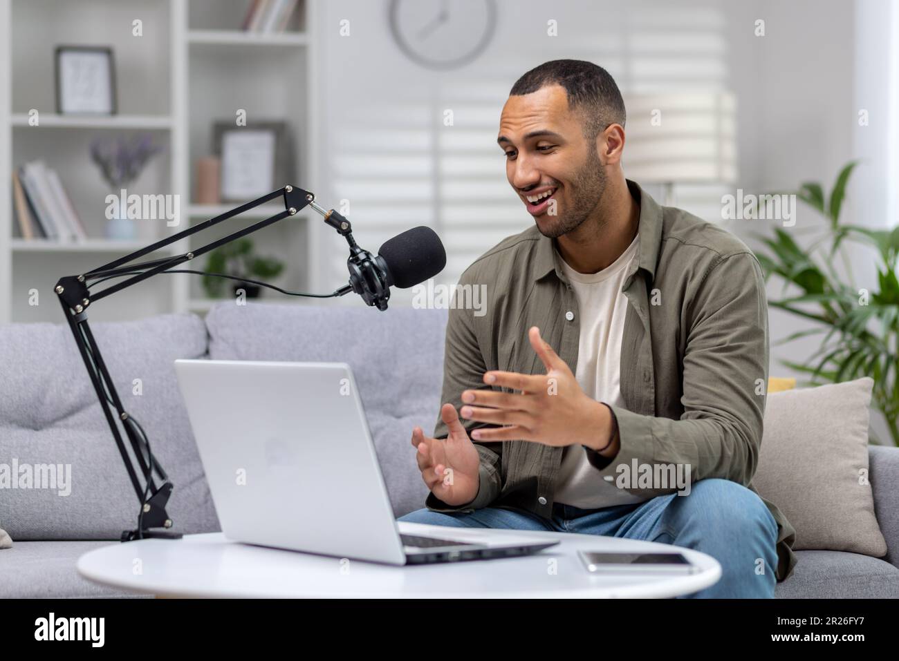 Jeune blogueur à la maison sur un canapé avec un ordinateur portable d'enregistrement en ligne podcast, homme avec le microphone professionnel en streaming depuis le salon, hispanique gaiement Banque D'Images