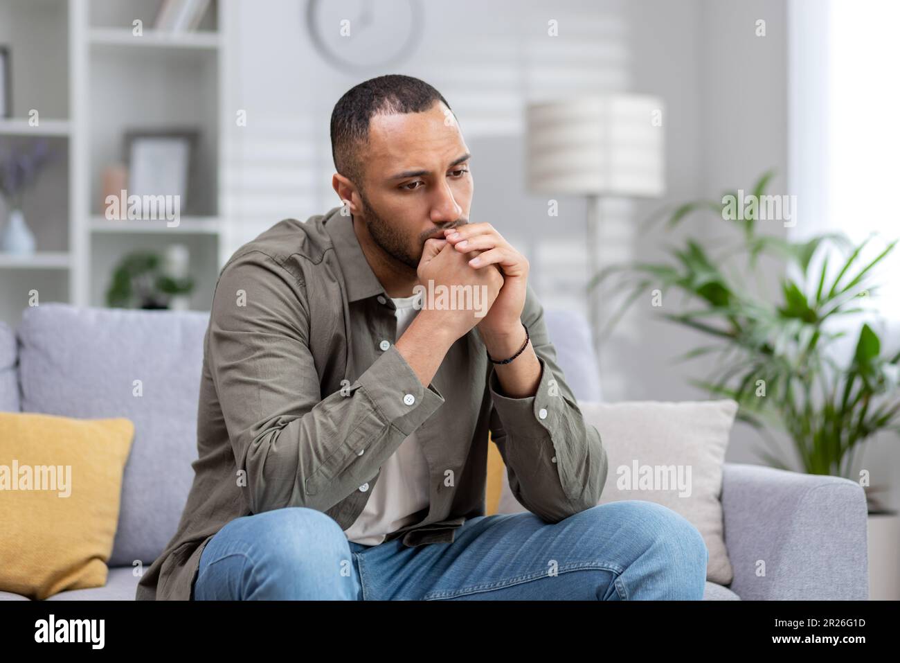 Homme contrarié assis seul à la maison, homme hispanique déprimé sur le canapé dans le salon dans des vêtements décontractés pensant à la décision difficile. Banque D'Images