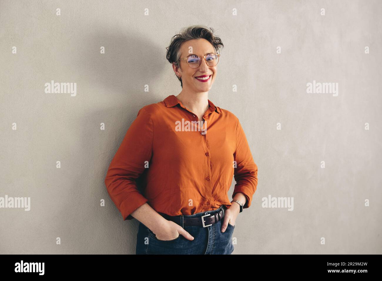 Portrait d'une femme d'affaires heureuse avec des lunettes souriant à l'appareil photo tout en se tenant contre un mur gris. Femme d'affaires mûre portant un casu d'affaires Banque D'Images