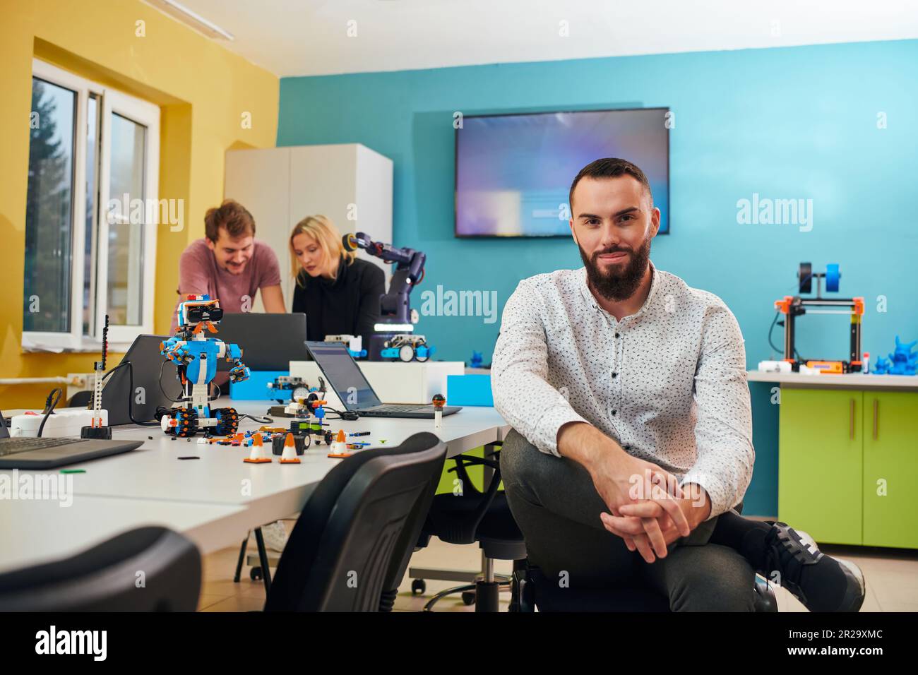 Un homme assis dans un laboratoire de robotique tandis que ses collègues en arrière-plan testent de nouvelles inventions robotiques de pointe. Banque D'Images