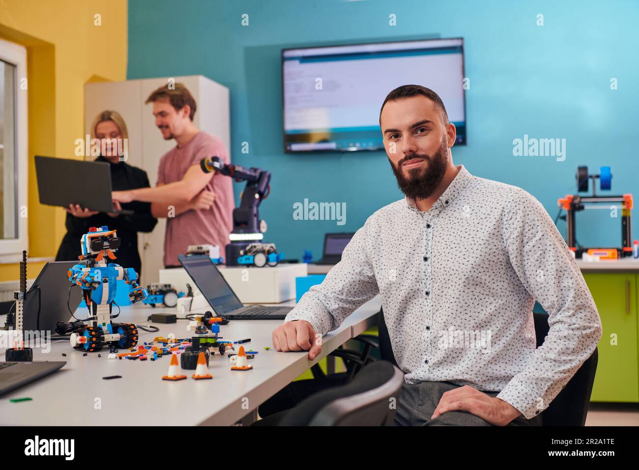 Un homme assis dans un laboratoire de robotique tandis que ses collègues en arrière-plan testent de nouvelles inventions robotiques de pointe. Banque D'Images