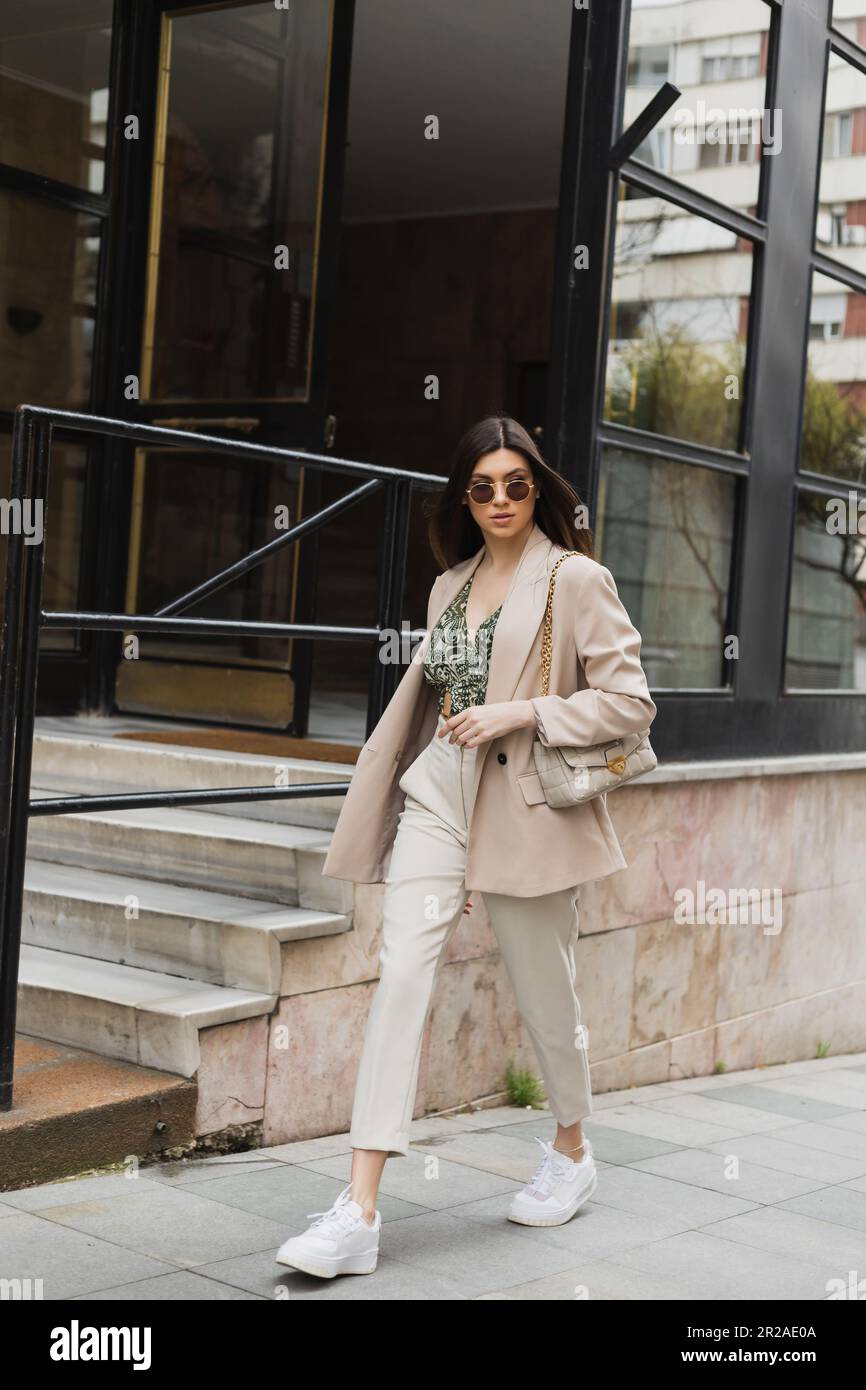 jeune femme en lunettes de soleil élégantes et tenue tendance avec pantalon  blanc et blazer beige marchant avec sac à main près du bâtiment moderne avec  fenêtres et escalier Photo Stock -