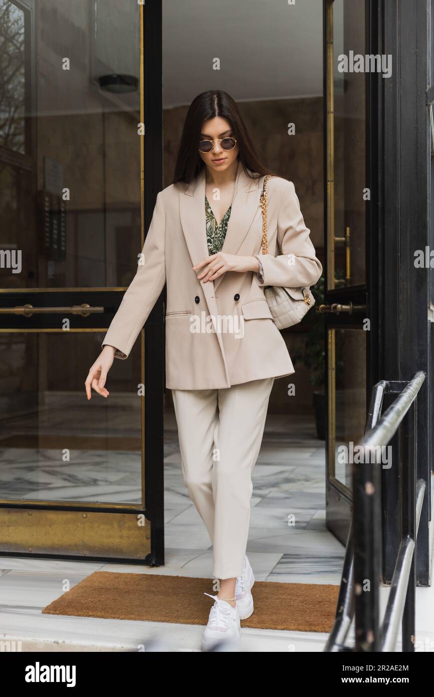 brunette et une jeune femme aux cheveux longs qui sortent du bâtiment moderne dans une tenue élégante et des lunettes de soleil tout en tenant un sac à main avec une sangle de chaîne sur le stre Banque D'Images