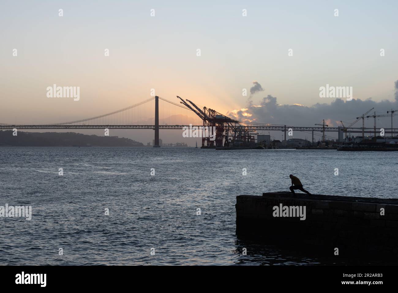 Une personne seule en silhouette debout sur la jetée dans le port de Lisbonne avec le pont du 25 avril et le terminal de fret en arrière-plan contre le coucher du soleil à Lisbonne Portu Banque D'Images