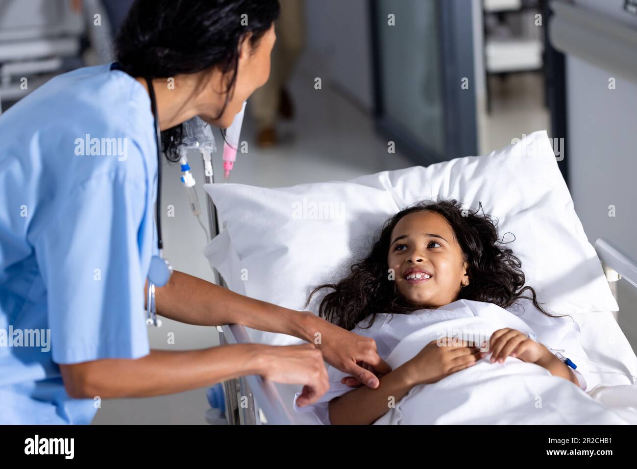 Femme biraciale médecin et fille souriante patiente dans le lit parlant dans le couloir à l'hôpital Banque D'Images