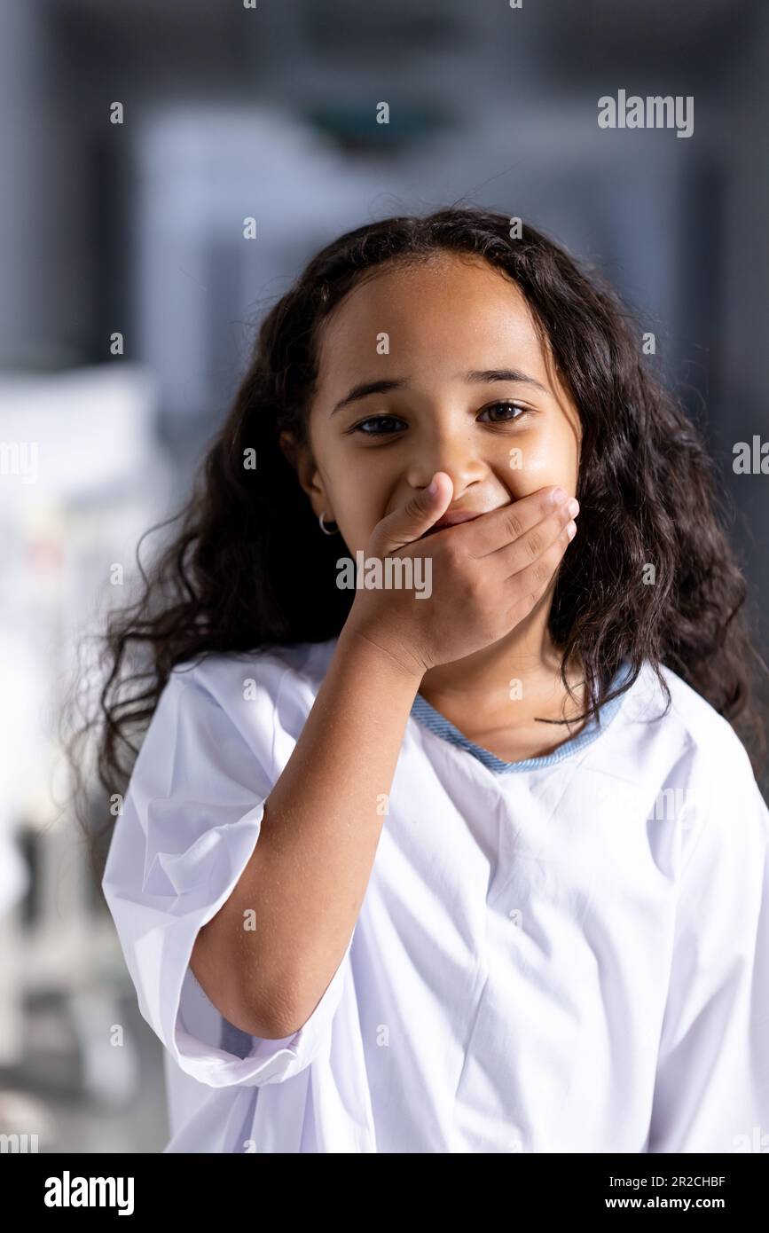 Portrait d'une jeune fille biraciale heureuse avec la main dans la bouche souriant dans le couloir de l'hôpital Banque D'Images