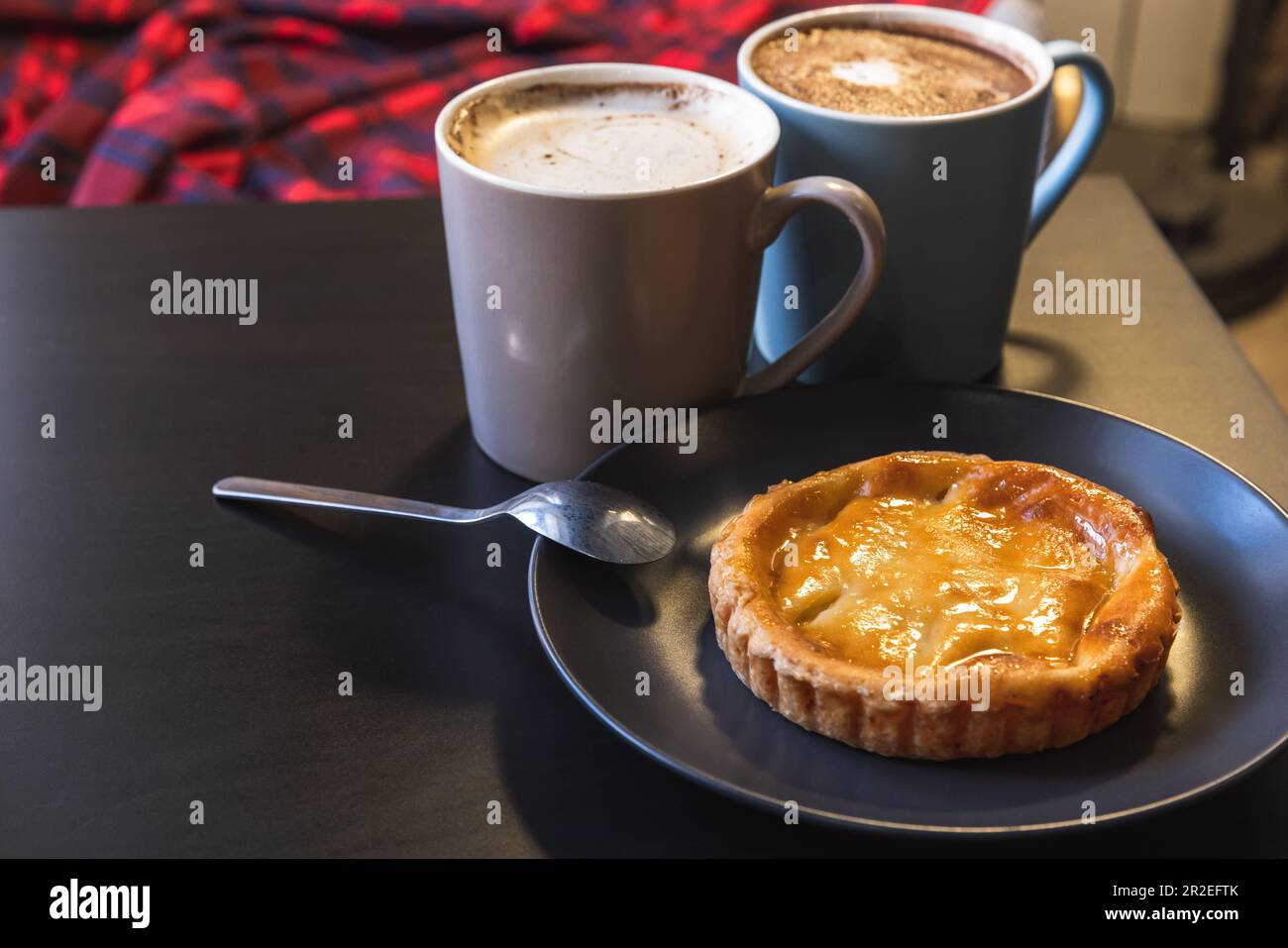 La pâte à tarte aux œufs portugais pastel de nata se trouve sur une table noire près de deux tasses de cappuccino Banque D'Images