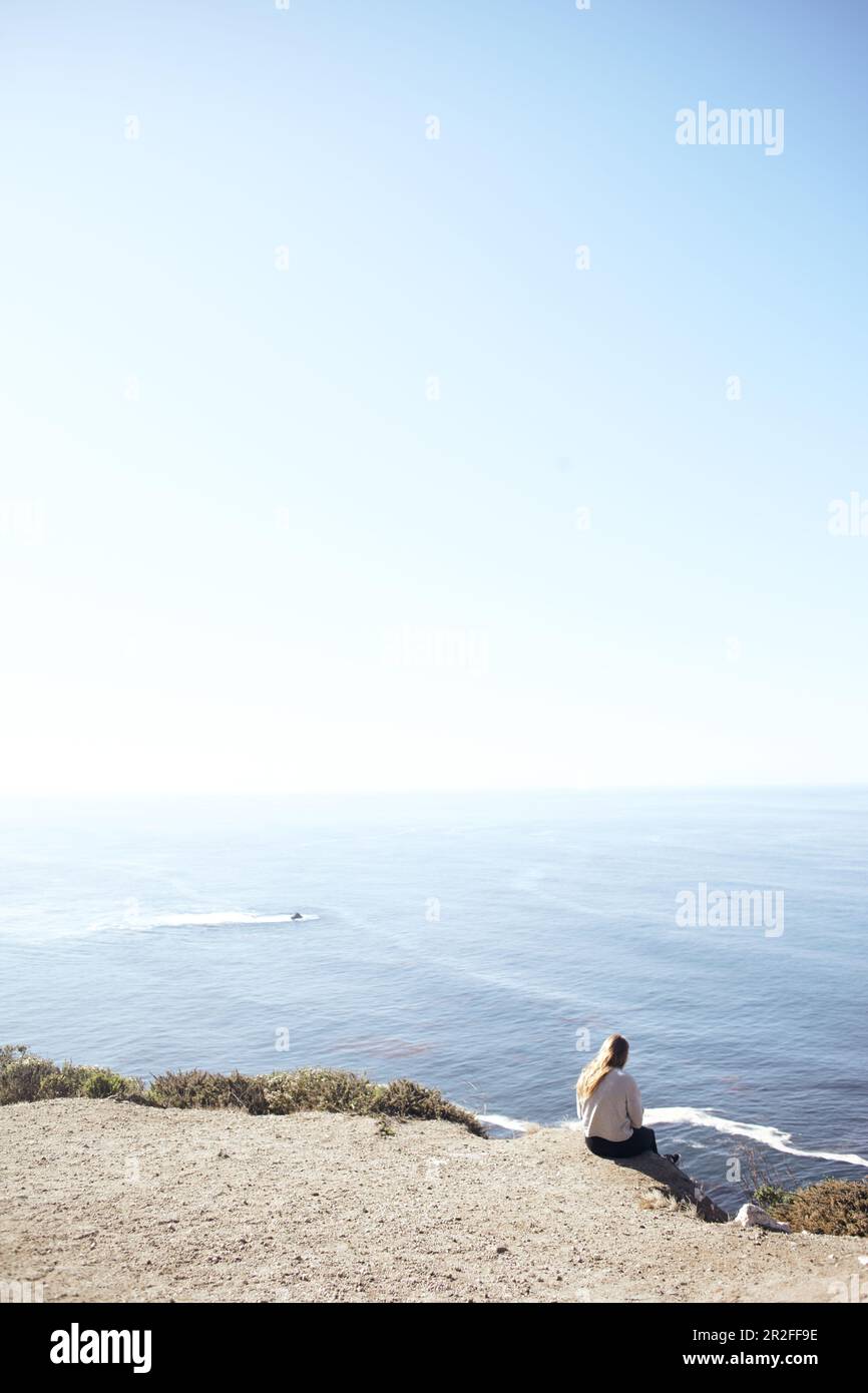 La jeune femme est assise sur une falaise et donne sur l'océan Pacifique, au bord de la Highway 1, Big sur State Park, Californie, États-Unis. Banque D'Images