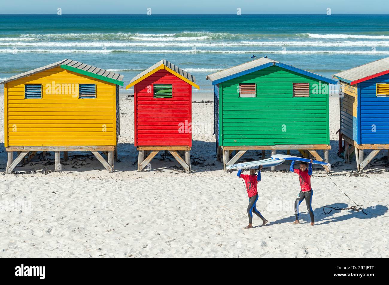 Des surfeurs souriants près des cabines de plage et des huttes de la plage de Muizenberg, un lieu de surf près du Cap, en Afrique du Sud. Banque D'Images