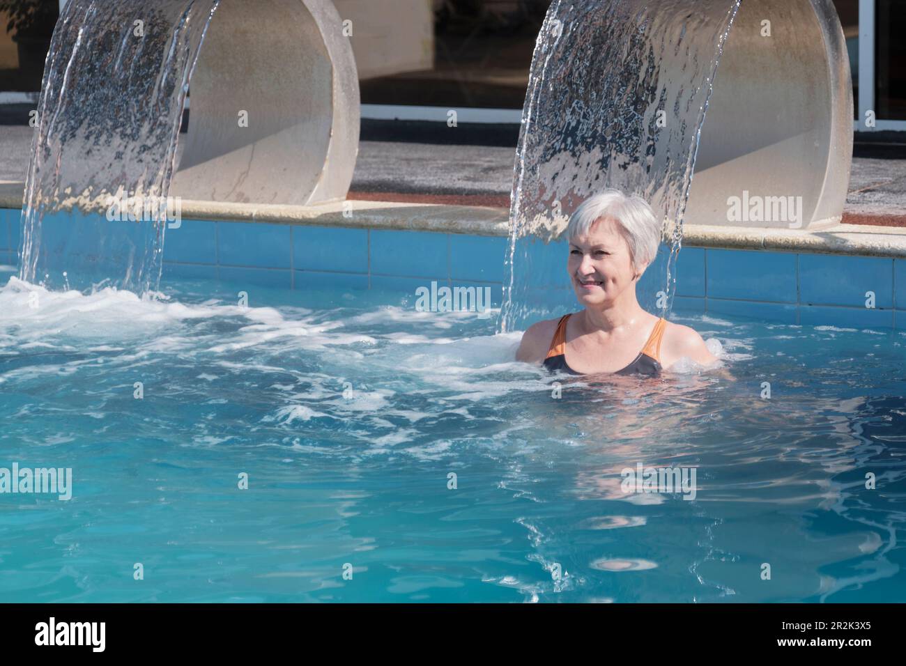 Femme caucasienne âgée qui se smilling avec des cheveux gris appréciant des jets d'eau tombant. Hydromassage dans la piscine thermale extérieure. Concept de personnes âgées actives. Banque D'Images