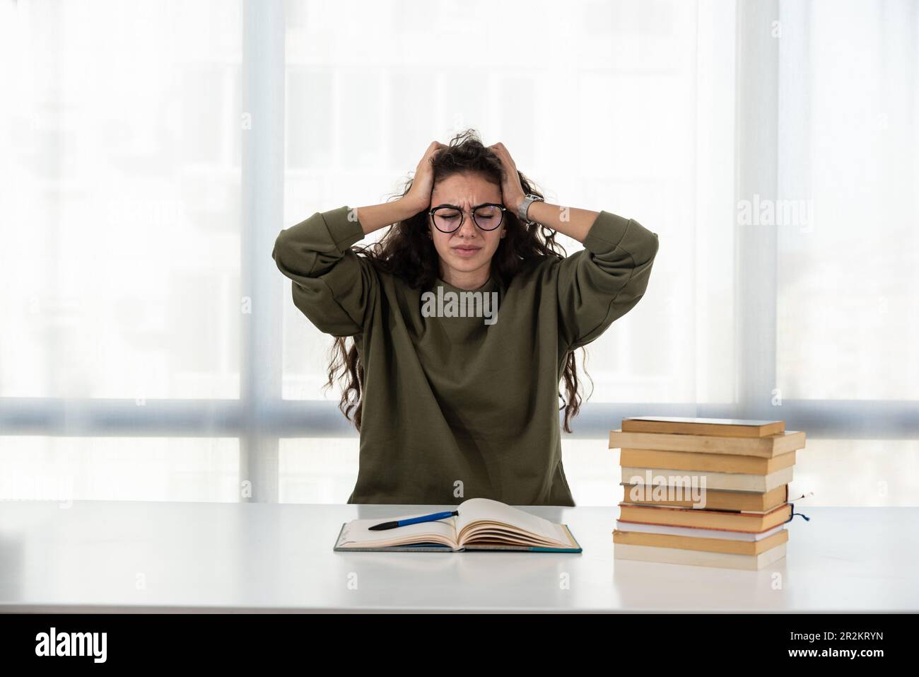 Surtravaillé. Une jeune femme fatiguée s'assoit au bureau de la maison bondé de livres cacher le visage dans les palmiers sentir mal de tête. Une femme exténuée se sent mal lo Banque D'Images