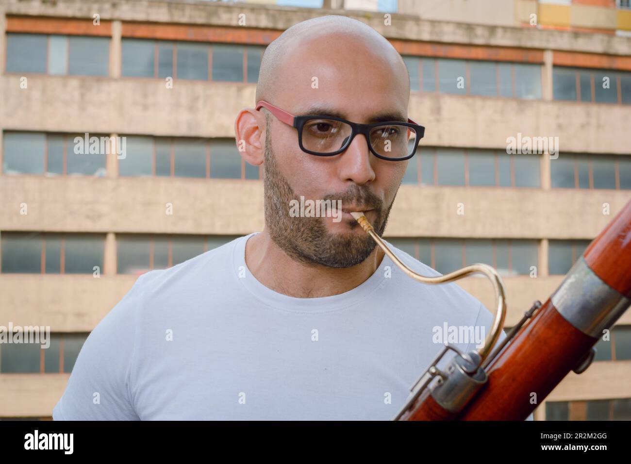 gros plan du jeune latino-homme avec des lunettes, une barbe et un chauve, portant un t-shirt blanc, jouant debout basson sur le balcon de l'appartement avec bui urbain Banque D'Images