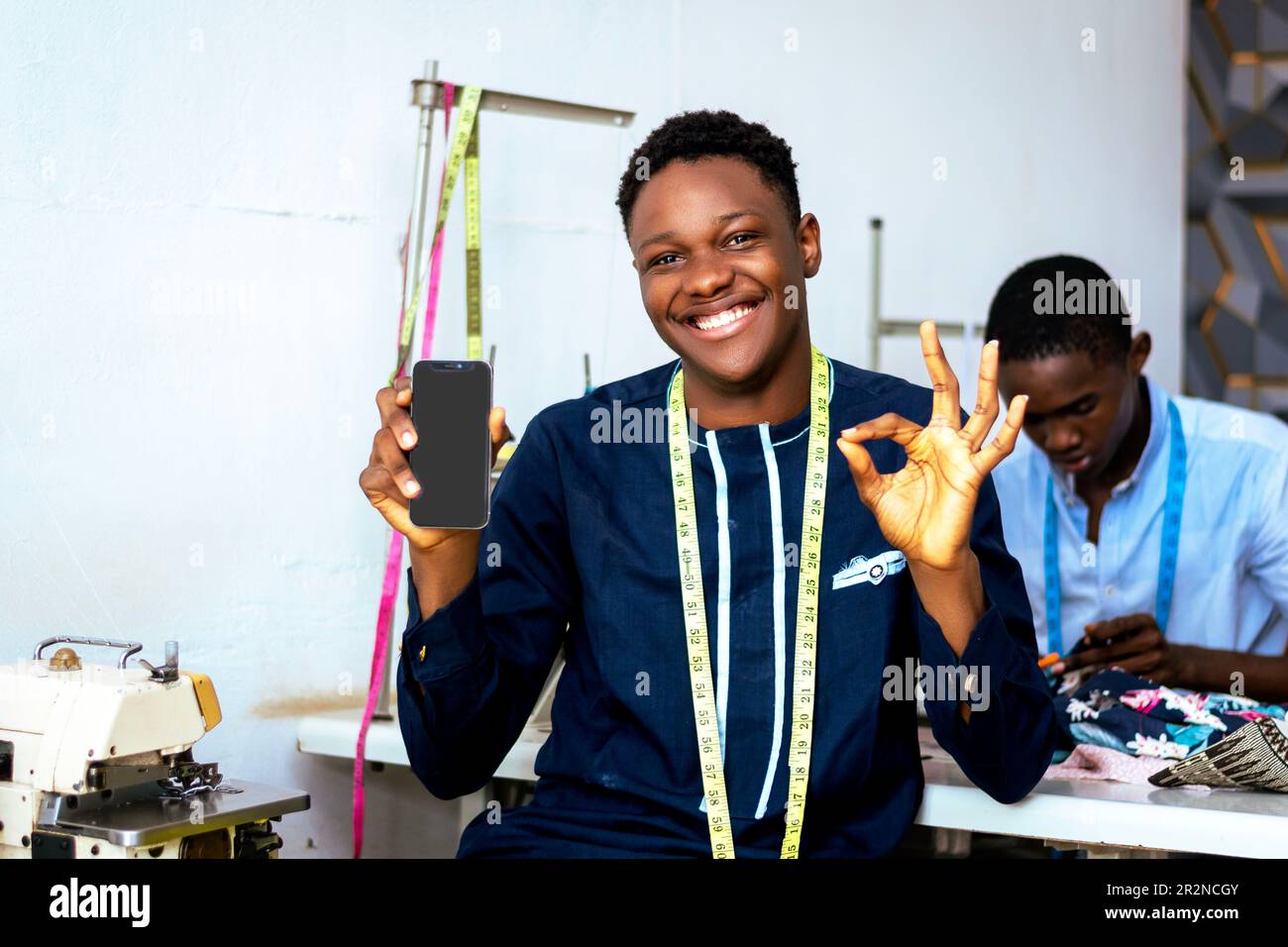 Un jeune designer de vêtements africains souriant tenant le téléphone et regardant l'appareil photo tout en étant isolé dans un environnement de magasin sur mesure Banque D'Images