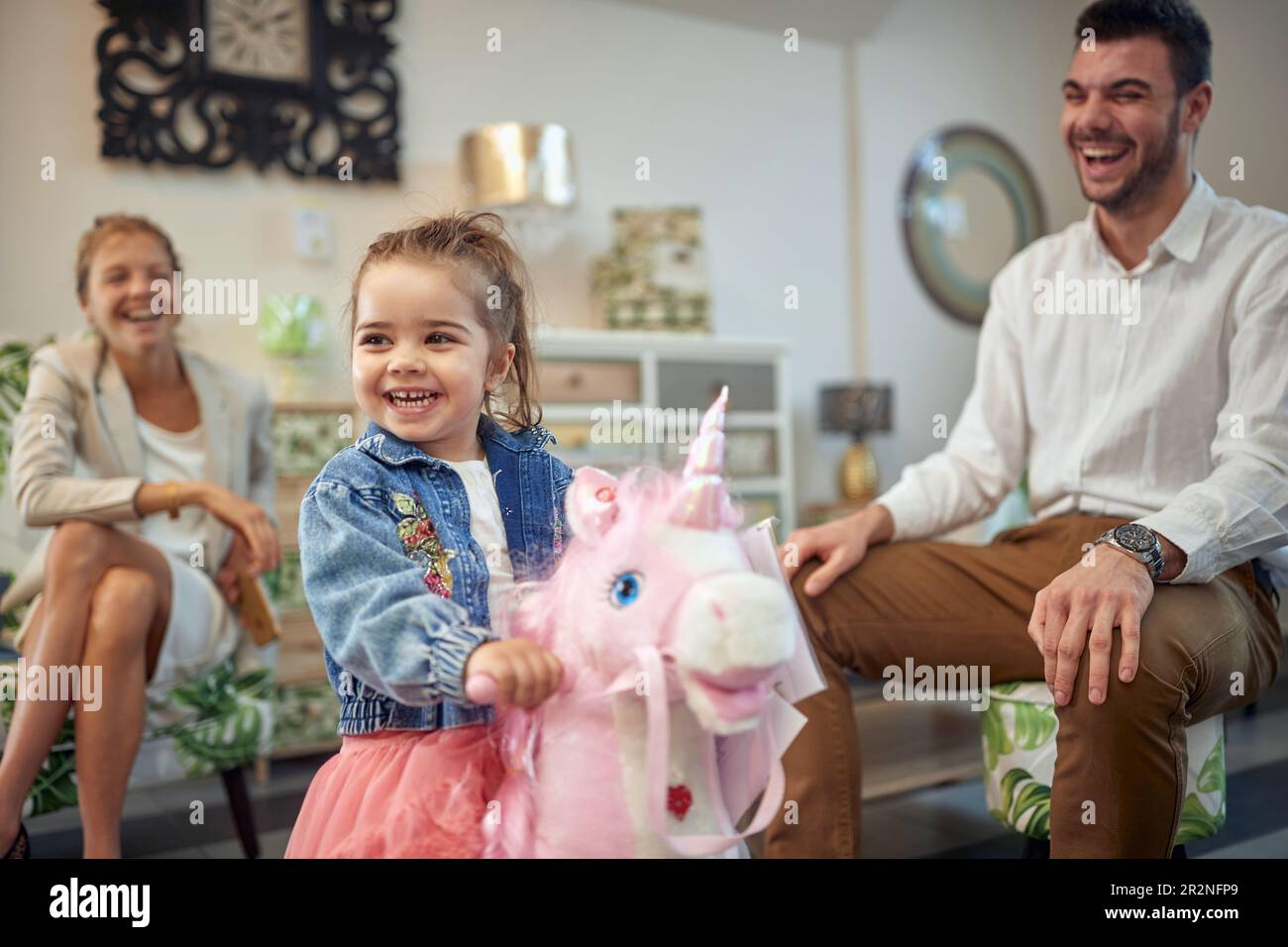 Adorable petite fille à cheval rose licorne dans une boutique de meubles avec ses parents assis à côté d'elle, riant de joie, se sentant heureux. S Banque D'Images