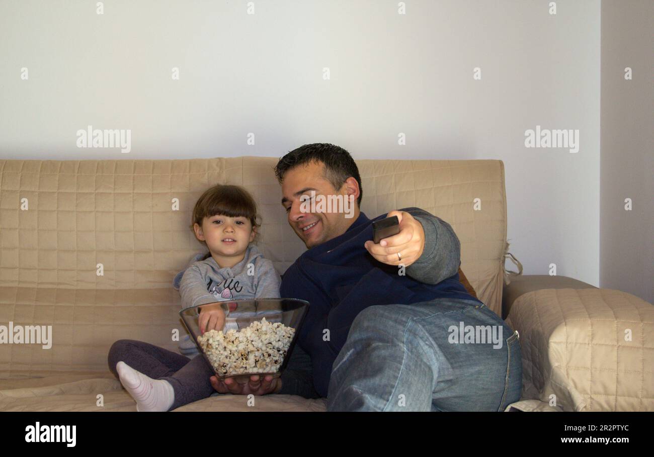 Image d'un père avec sa fille allongée sur le canapé à la maison tout en regardant la télévision et en mangeant du pop-corn. Banque D'Images