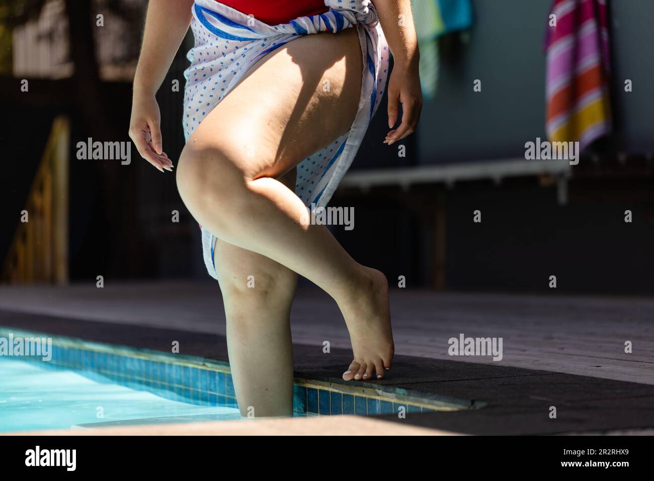 Partie basse de la femme biraciale non modifiée en maillot de bain rouge et serviette de bain entrant dans la piscine. Été, temps libre, détente et vacances. Banque D'Images
