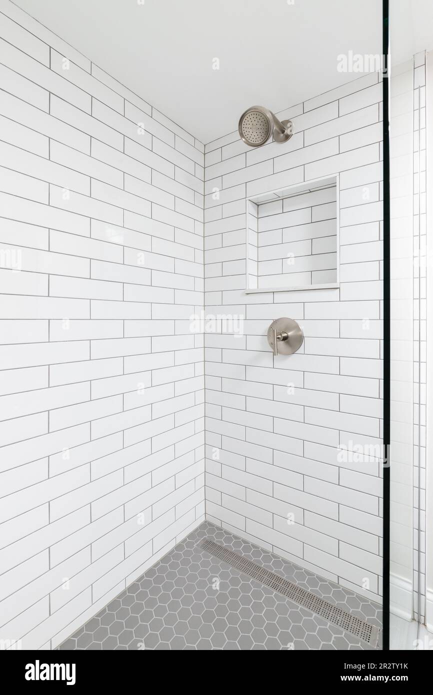 Une douche de salle de bains avec des murs de carreaux de métro blancs, une étagère encastrée, une pomme de douche en bronze et un sol carrelé hexagonal gris. Banque D'Images