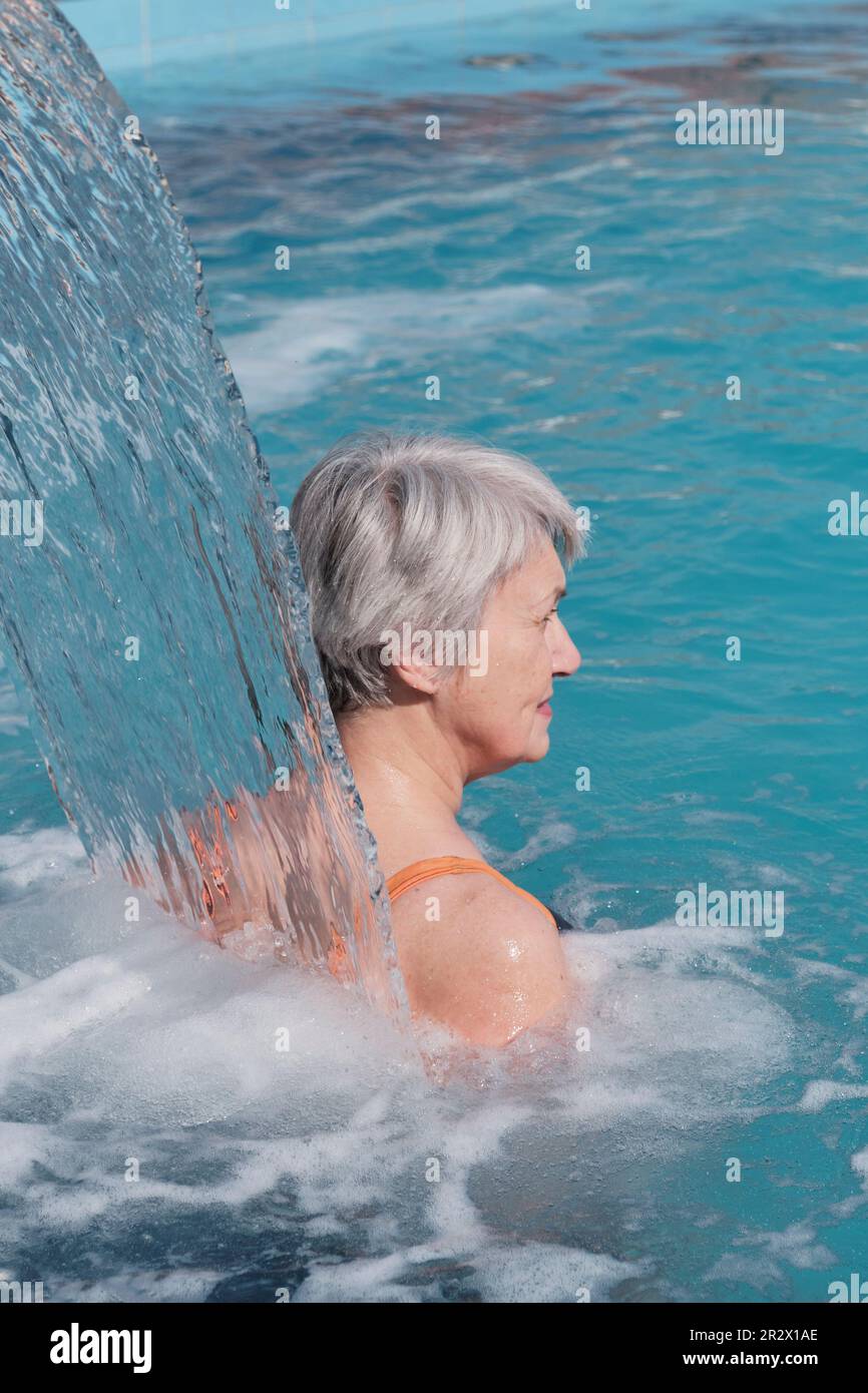 Femme âgée avec des cheveux gris près de la chute du flux d'eau. Vue arrière. Hydromassage dans la piscine thermale extérieure. Concept de personnes âgées actives. Banque D'Images