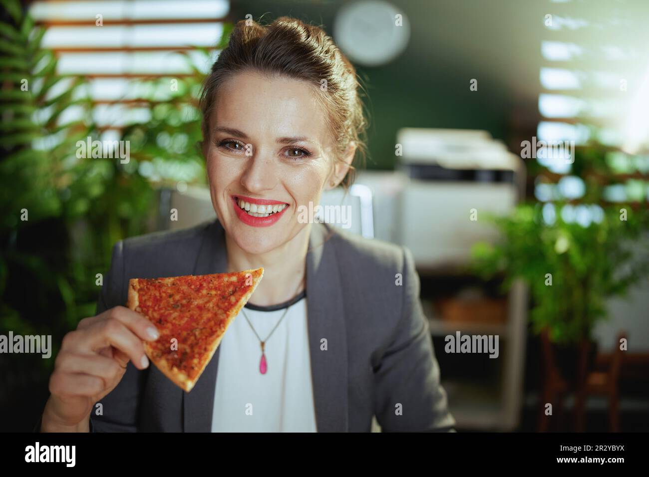Un lieu de travail durable. femme moderne souriante dans un costume d'affaires gris dans un bureau moderne vert avec pizza. Banque D'Images