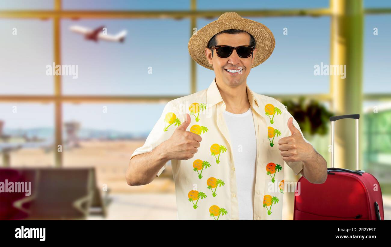 Latin homme en vacances portant une chemise à fleurs chapeau lunettes de soleil à l'aéroport faisant des pouces heureux vers le haut geste avec les mains. Approbation de l'expression en regardant la came Banque D'Images
