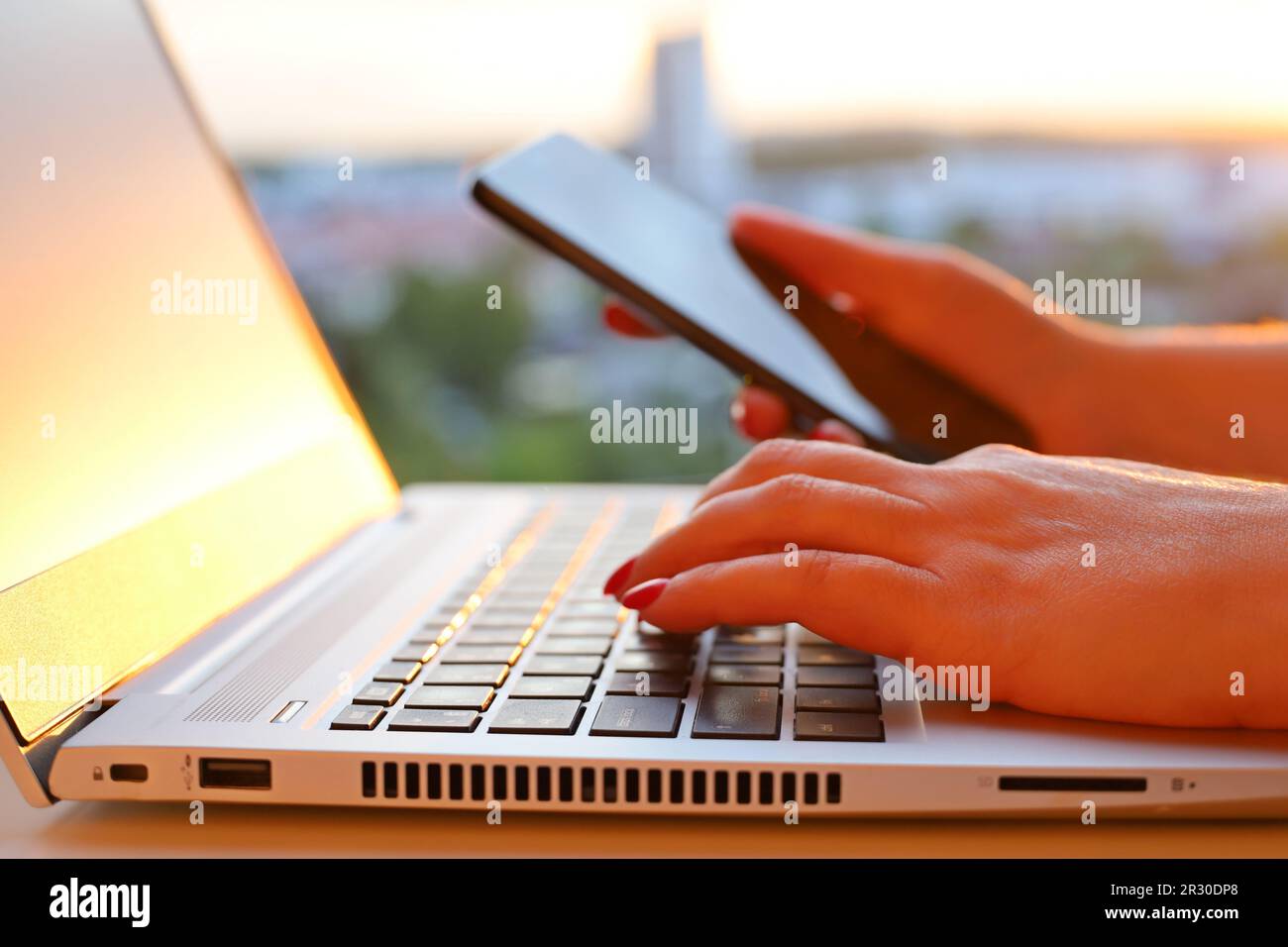 Femme avec un smartphone assis au clavier de l'ordinateur portable près de la fenêtre au coucher du soleil. Espace de travail confortable, concept de communication en ligne, bureau ou travail à domicile Banque D'Images