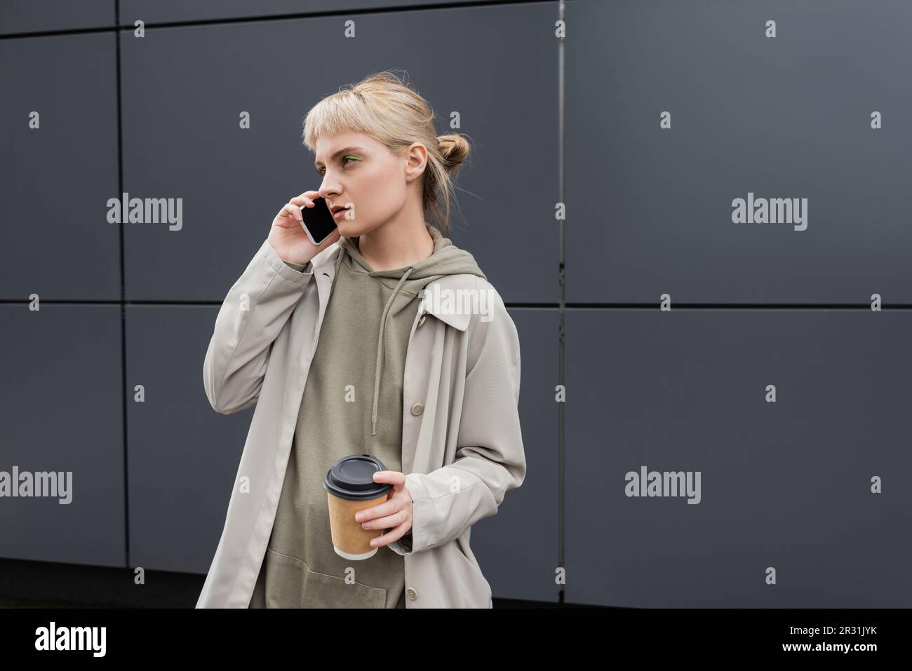 jeune femme élégante avec des langes et des cheveux blonds tenant une tasse en papier avec du café à emporter tout en parlant sur un smartphone et debout dans un sweat à capuche et un manteau près de gr Banque D'Images