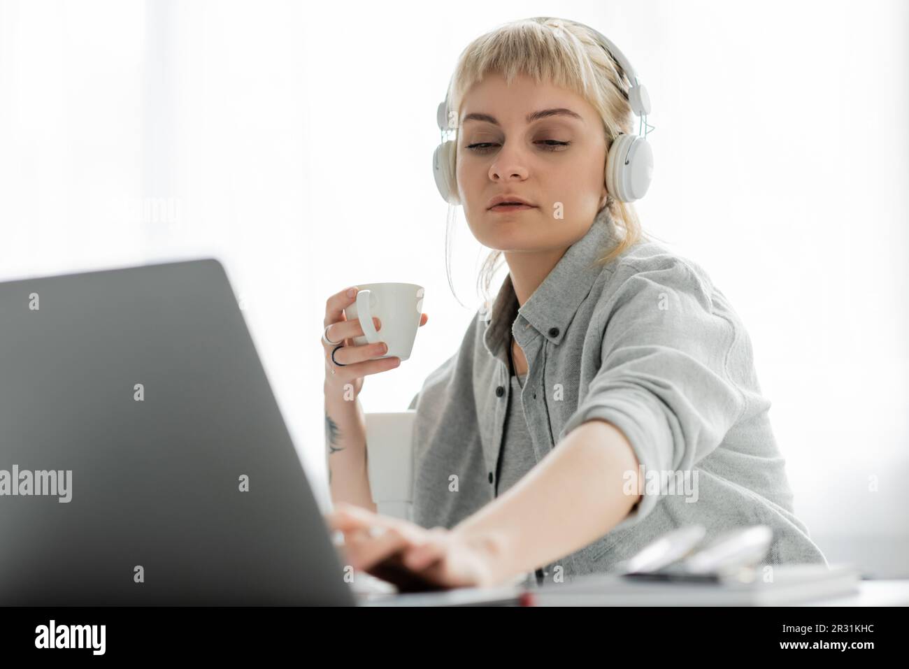 jeune freelance avec cheveux blonds, bangs et tatouage à la main assis dans un casque sans fil et tenant une tasse de café près d'un ordinateur portable et un bloc-notes flou Banque D'Images