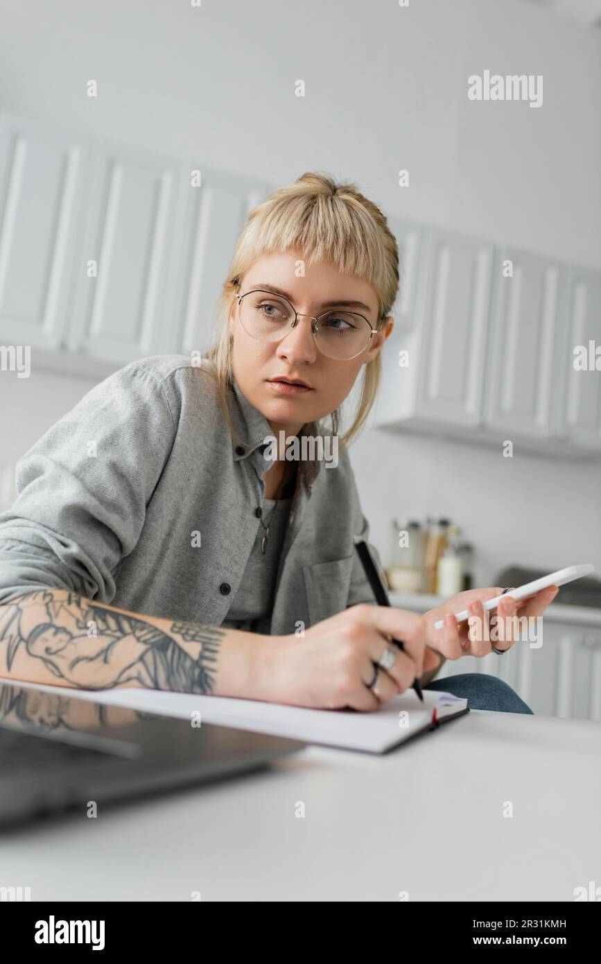 jeune femme avec tatouage à la main écrire dans un carnet, prendre des notes, tenir un smartphone et un stylo près d'un ordinateur portable sur une table blanche, avant-plan flou, travail fro Banque D'Images