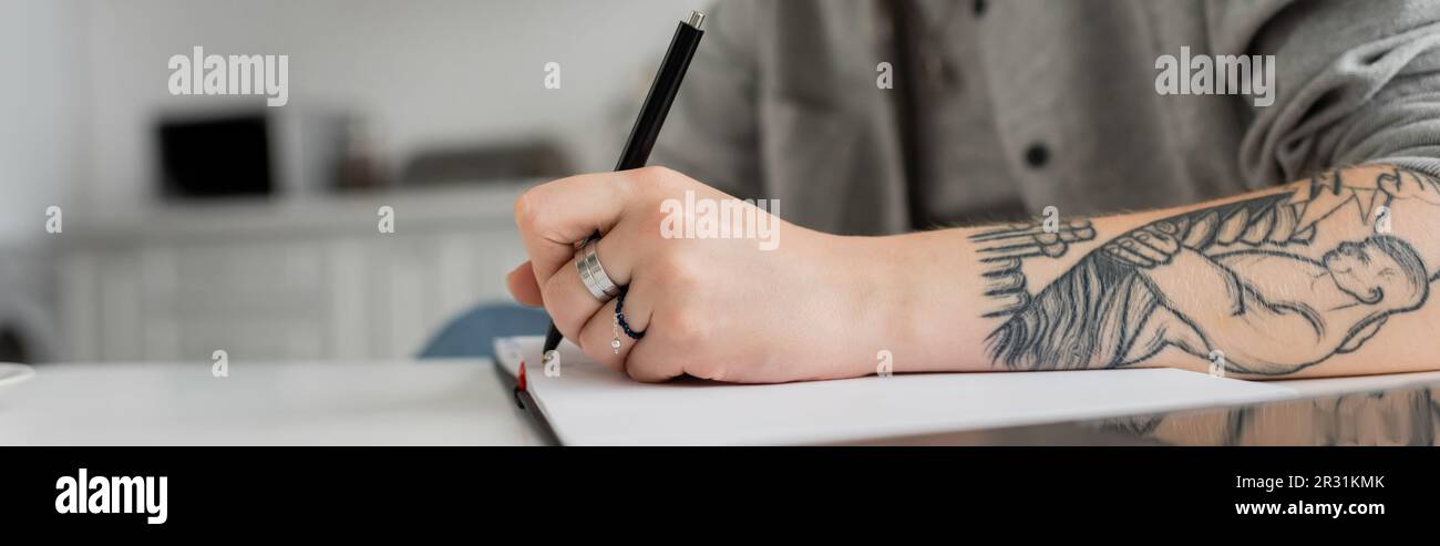 prise de vue rognée d'une jeune femme avec tatouage à la main, écriture dans un carnet, prise de notes, inspiration tout en tenant le stylo près de l'ordinateur portable sur une table blanche, bl Banque D'Images