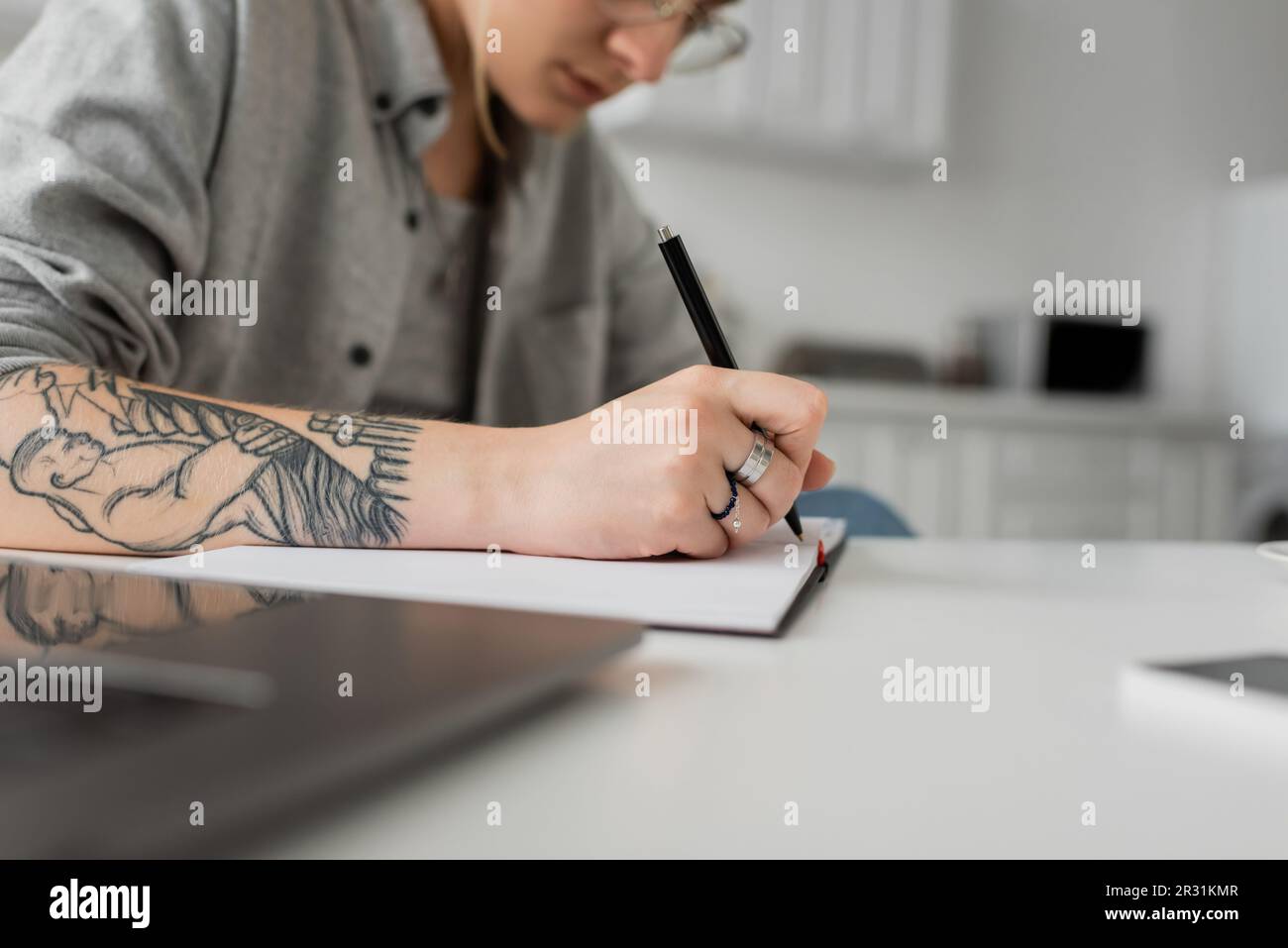 vue rognée de la jeune femme avec tatouage à la main écrire dans le carnet, prendre des notes, ayant l'inspiration tout en tenant le stylo près de l'ordinateur portable sur une table blanche, bl Banque D'Images