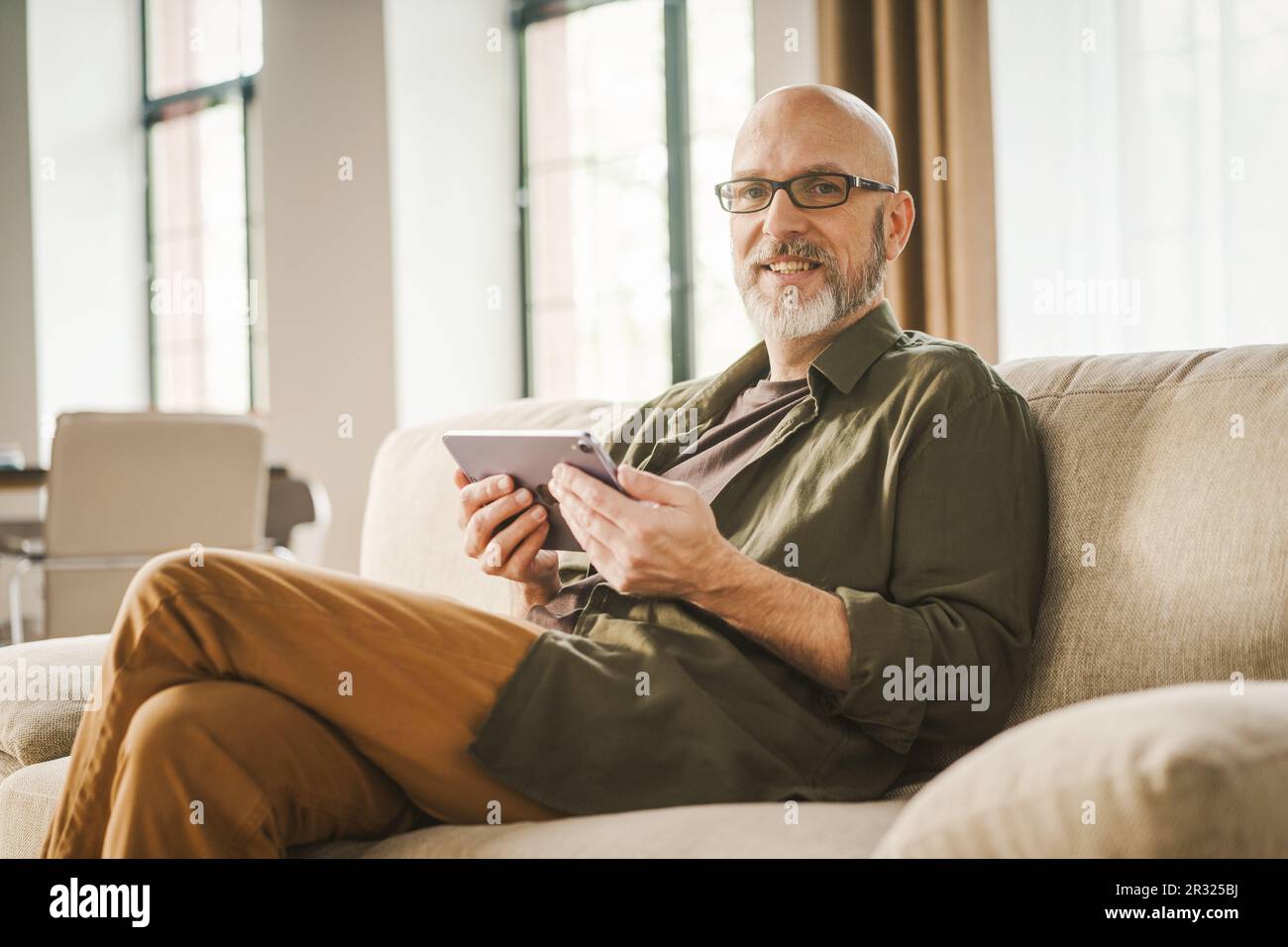 Un homme expérimenté heureux passe du temps à la maison, profitant de la commodité de la technologie moderne. Il s'est assis sur un canapé, a pris l'enrosé en regardant des services en ligne sur un Tablet PC. . Photo de haute qualité Banque D'Images