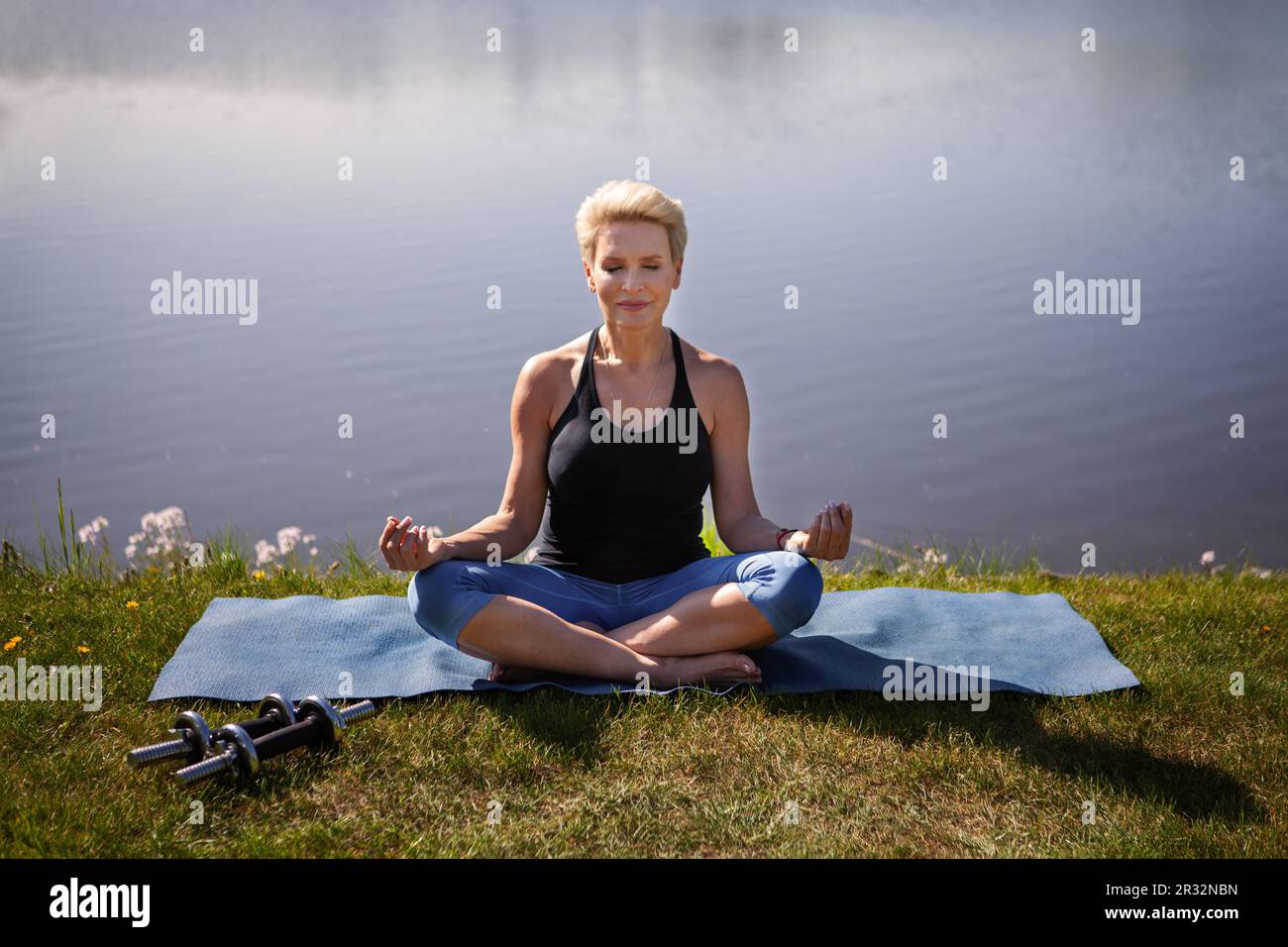 Paisible femme mature pratique le yoga à l'extérieur. Athlète féminine assise en position lotus sur fond d'eau de la nature. Un style de vie actif et de la santé Banque D'Images