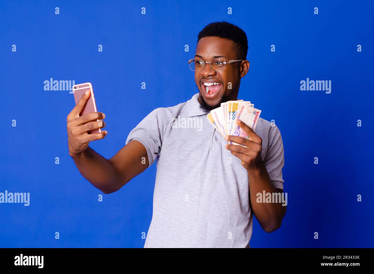 Jeune homme africain charmant sur des lunettes prenant selfie avec son téléphone portable et tenant de l'argent, sur un fond isolé Banque D'Images