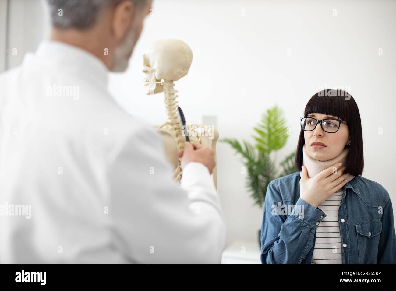 Vue sur l'épaule d'une belle jeune femme portant un étrésillon de cou et plantant sur le squelette humain utilisé par l'homme mature. Femme inquiète dans les lunettes rendant visite à une personne médicale après une lésion de la colonne vertébrale pour consultation. Banque D'Images