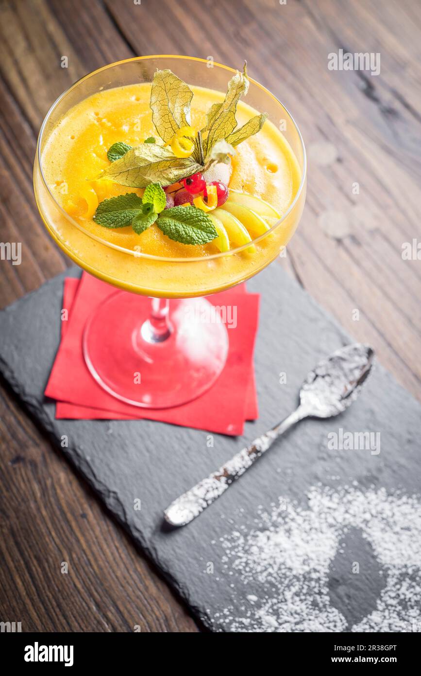Sorbet mangue décoré de zeste d'agrumes, de menthe, de groseille dans un verre avec une serviette rouge et une cuillère sur un panneau d'ardoise et un fond en bois Banque D'Images