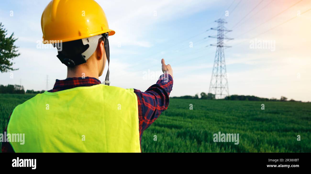 Ouvrier de la construction pointant vers le pylône d'électricité. Électricien portant un casque de sécurité jaune et un gilet fluorescent sur le terrain, zone de travail. OSH. Banque D'Images