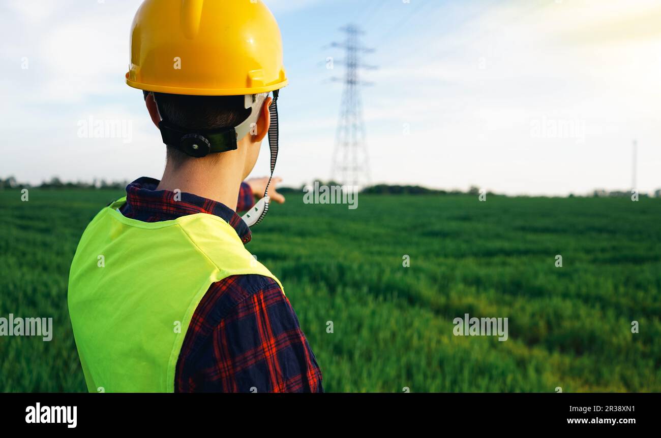 Ouvrier de la construction pointant vers le pylône d'électricité. Électricien portant un casque de sécurité jaune et un gilet fluorescent sur le terrain, zone de travail. OSH. Banque D'Images