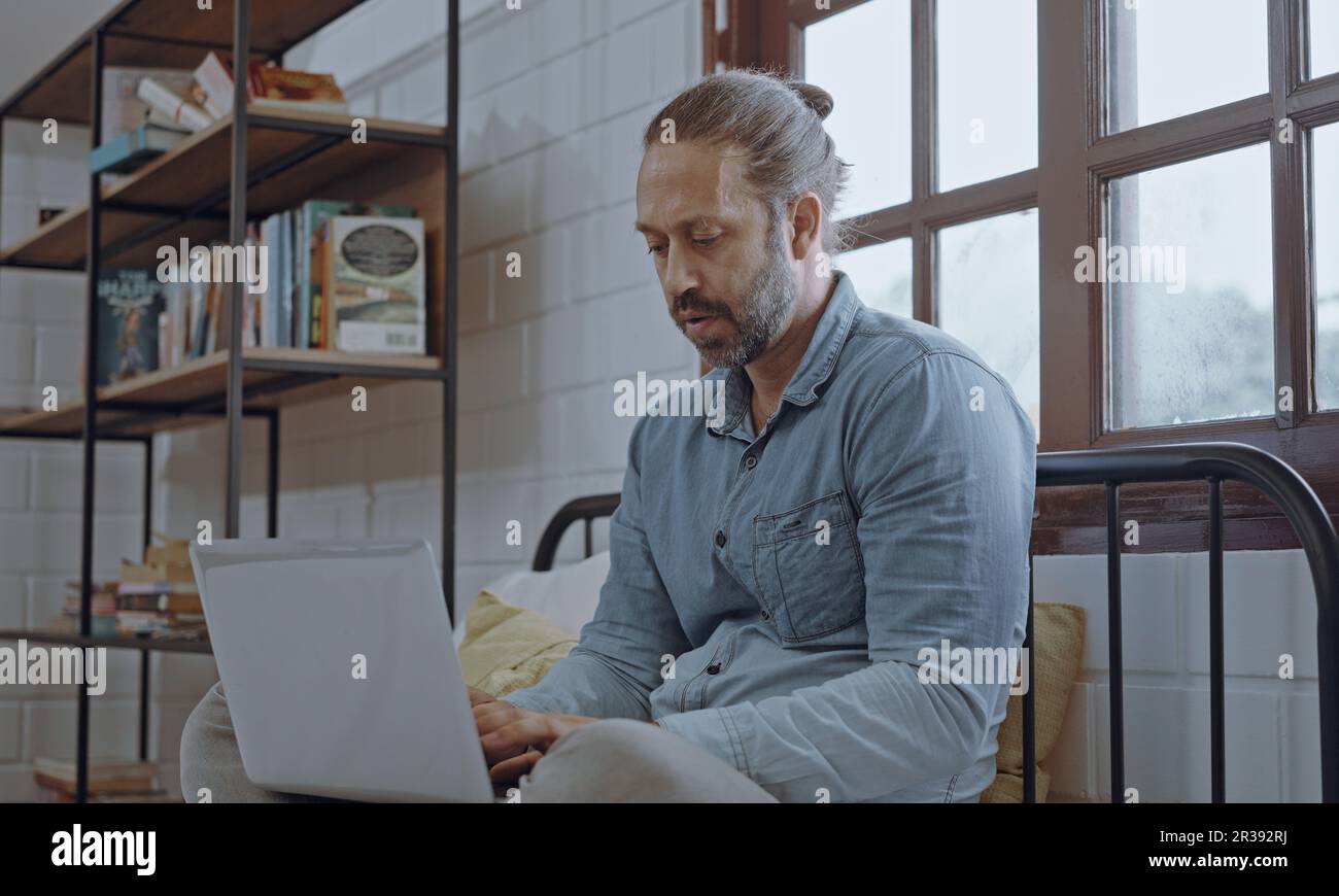 Un jeune homme inquiet à l'aide d'un ordinateur portable assis sur le lit dans la chambre. Banque D'Images